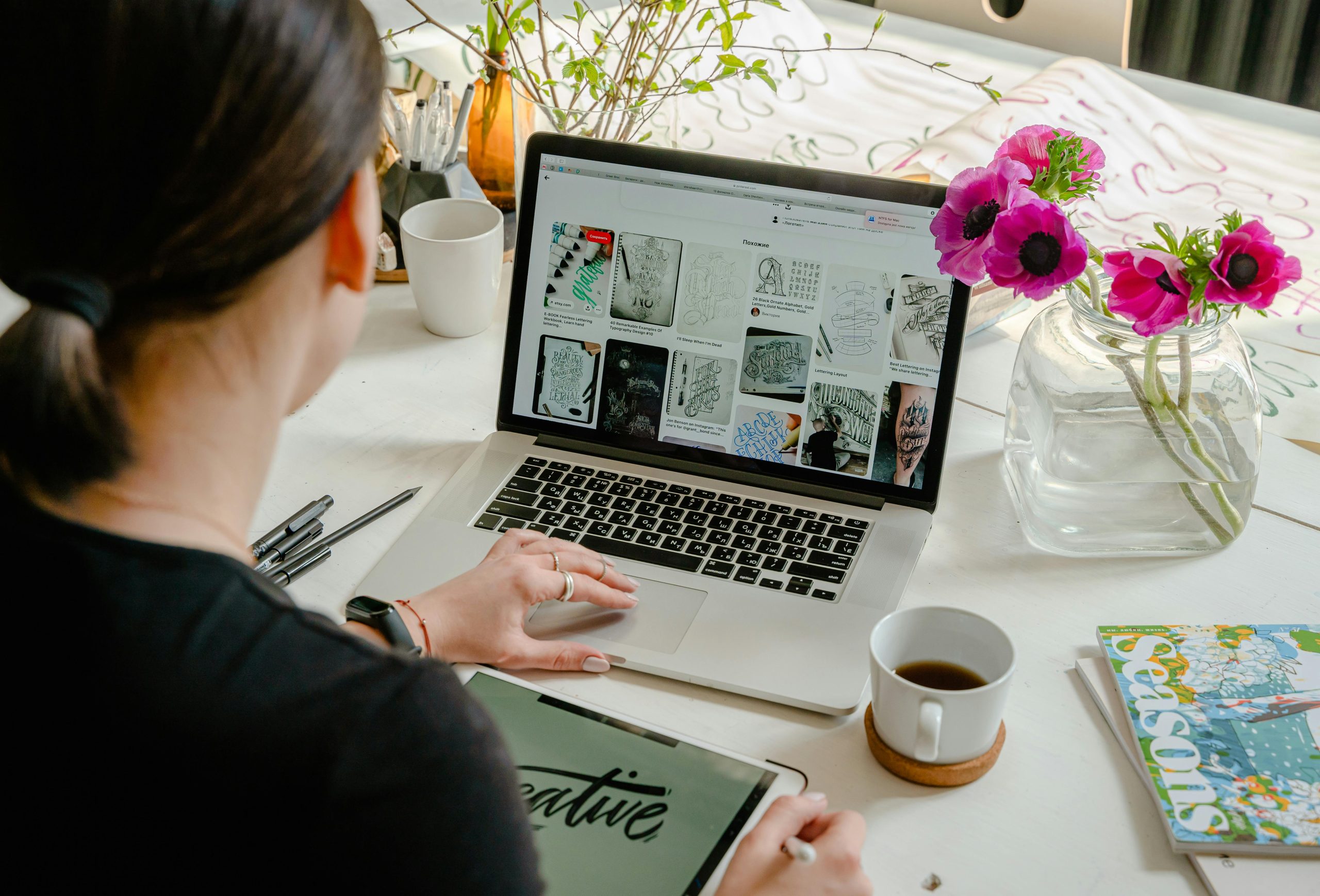 A female creative designer sitting at desk with laptop and notebooks with CREATIVE on it, and coffee mug. She is creating recurring revenue packages for her clients.