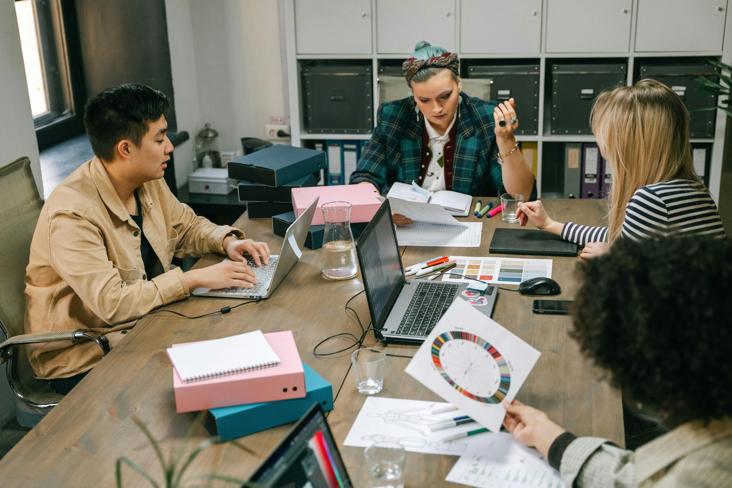 Creative agency team collaborating at desk with laptops, color samples, and project materials — managing money and project costs for subcontracted work.