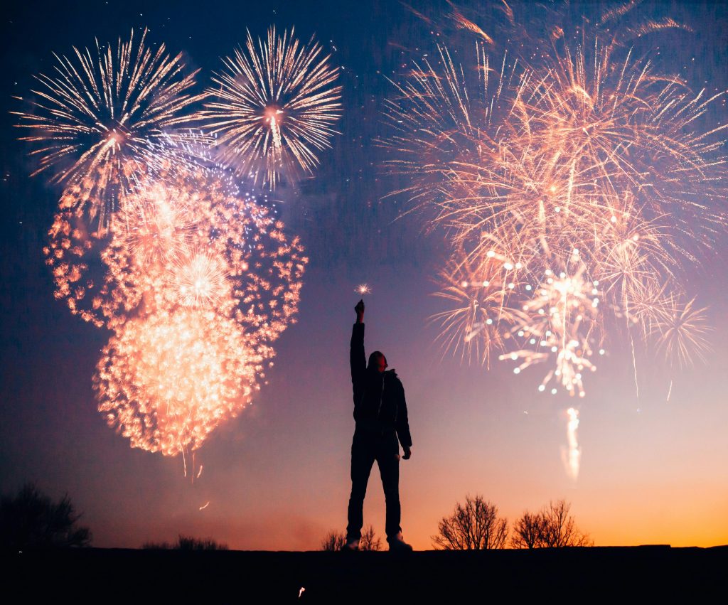 A person holding a sparkler stands against a sunset sky as fireworks explode above them, symbolizing creative freedom and personal empowerment.